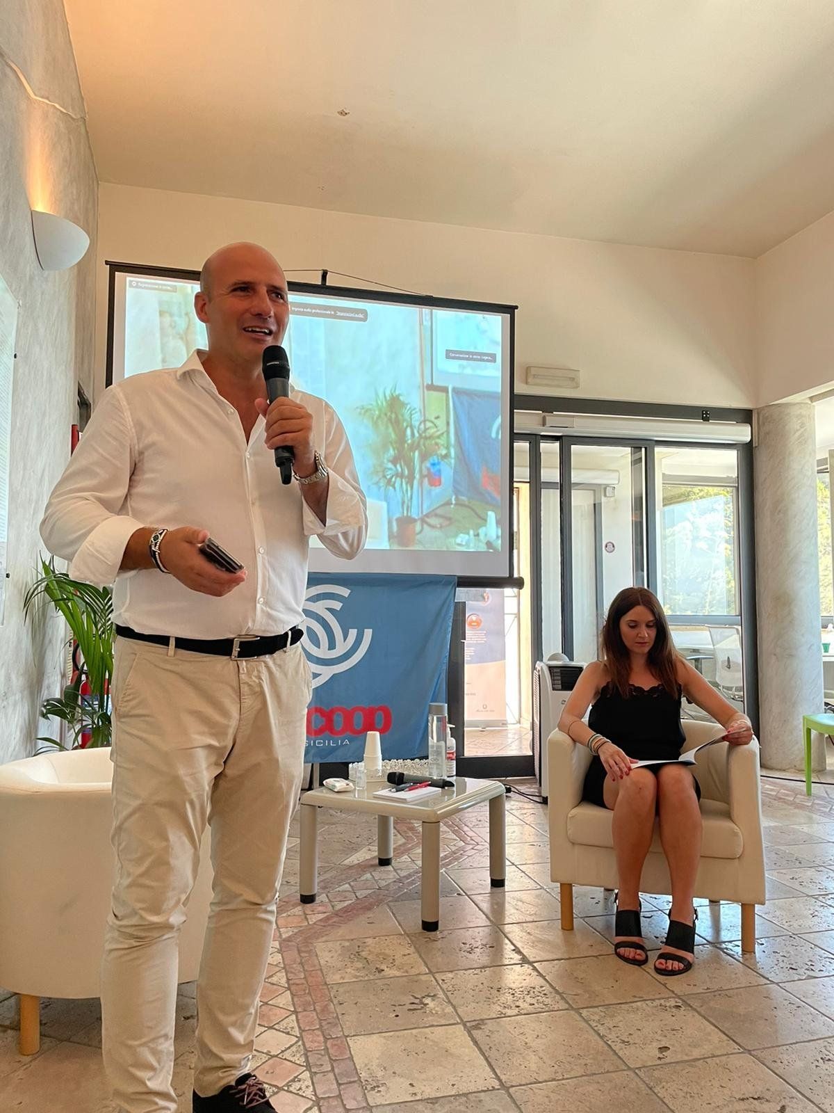 Un uomo è in piedi davanti a un microfono mentre una donna è seduta su una sedia.