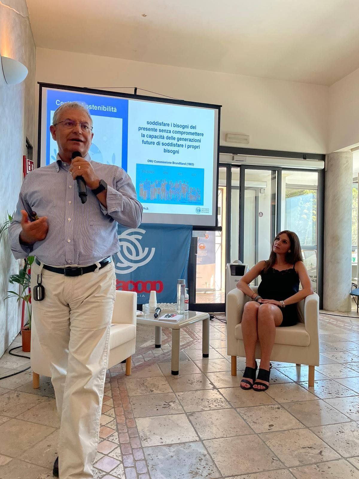 Un uomo parla in un microfono mentre una donna è seduta su una sedia.