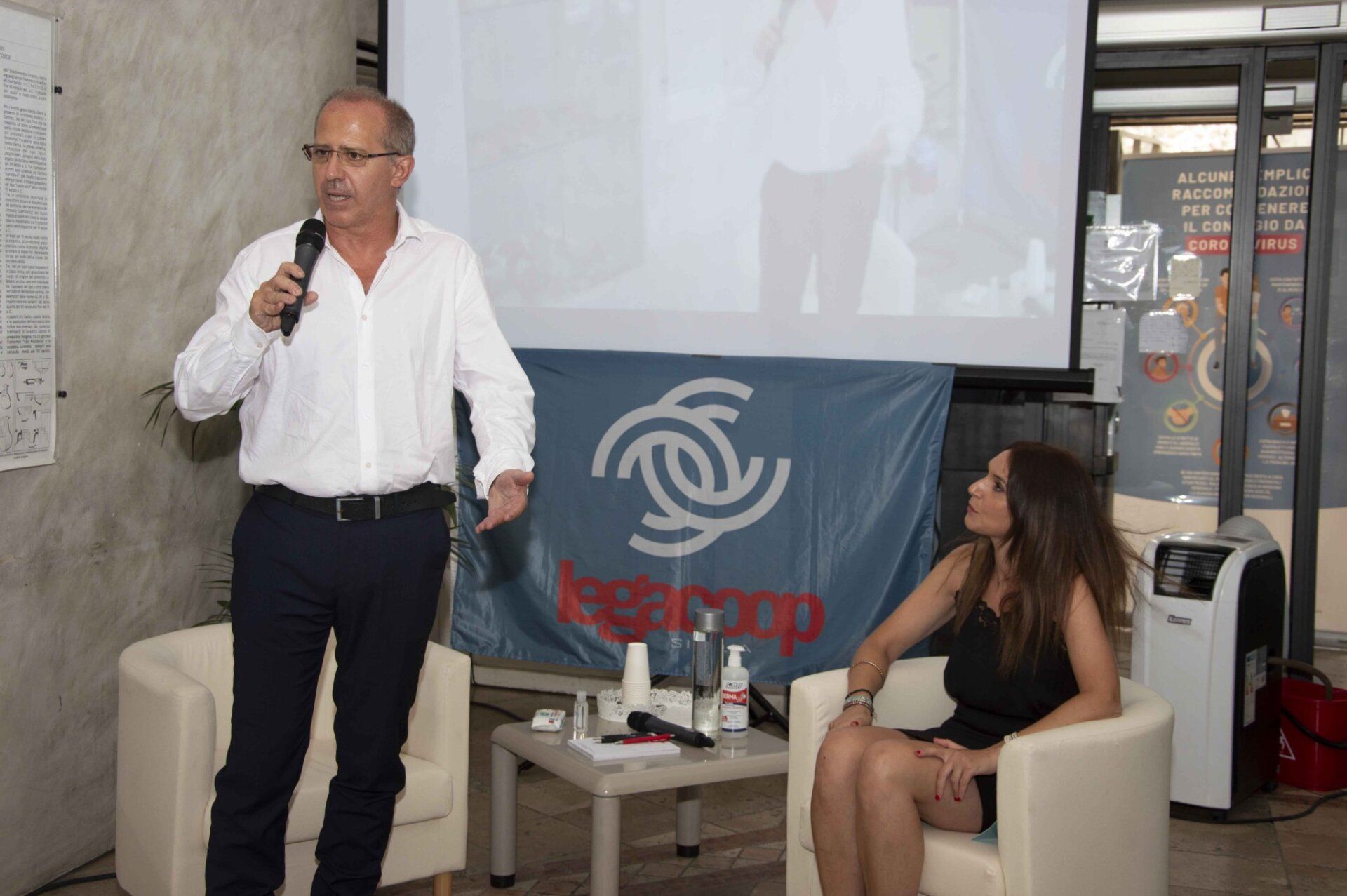 Un uomo parla al microfono mentre una donna è seduta su una sedia.