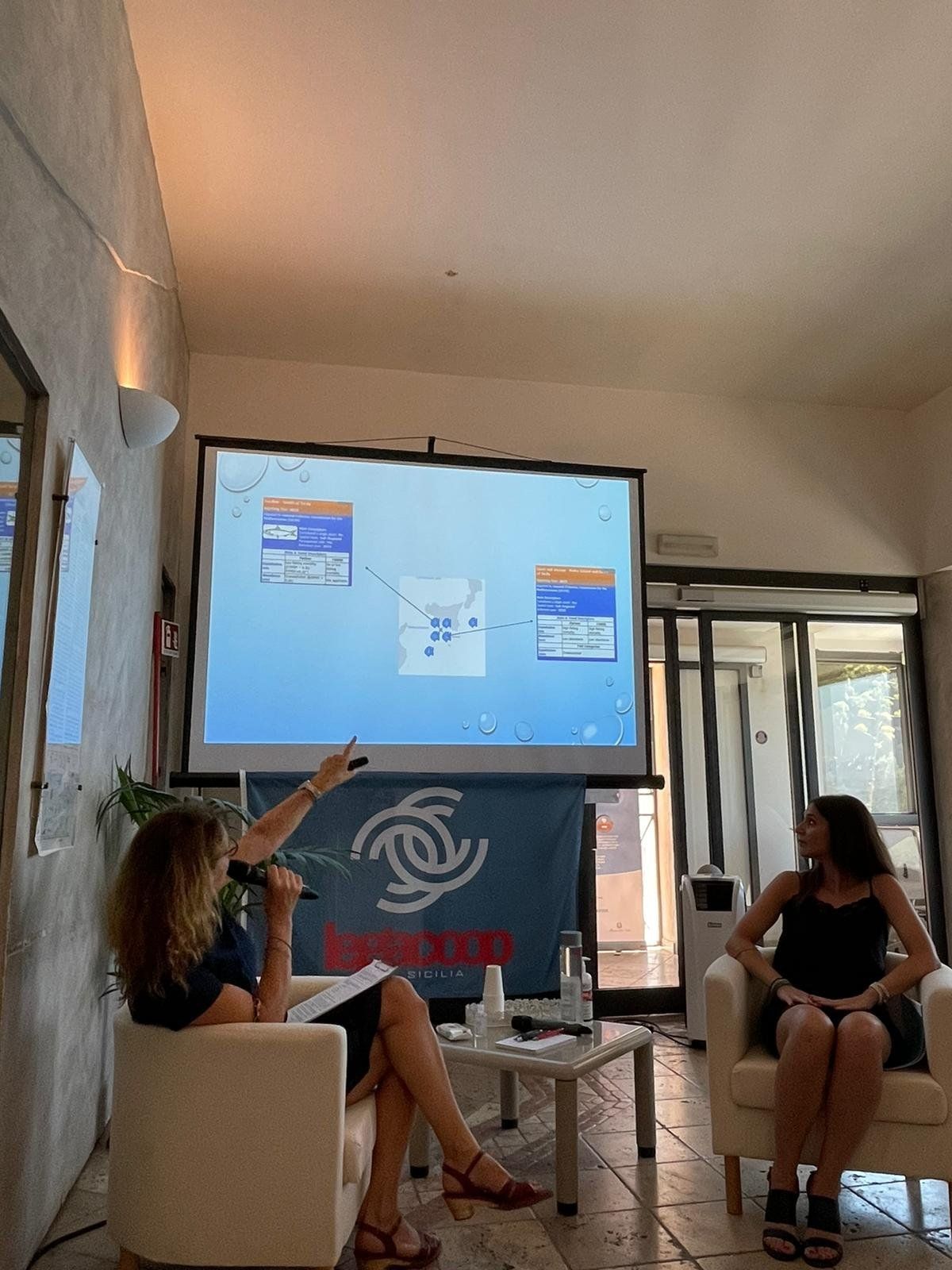 Due donne sono sedute su delle sedie davanti allo schermo di un proiettore.