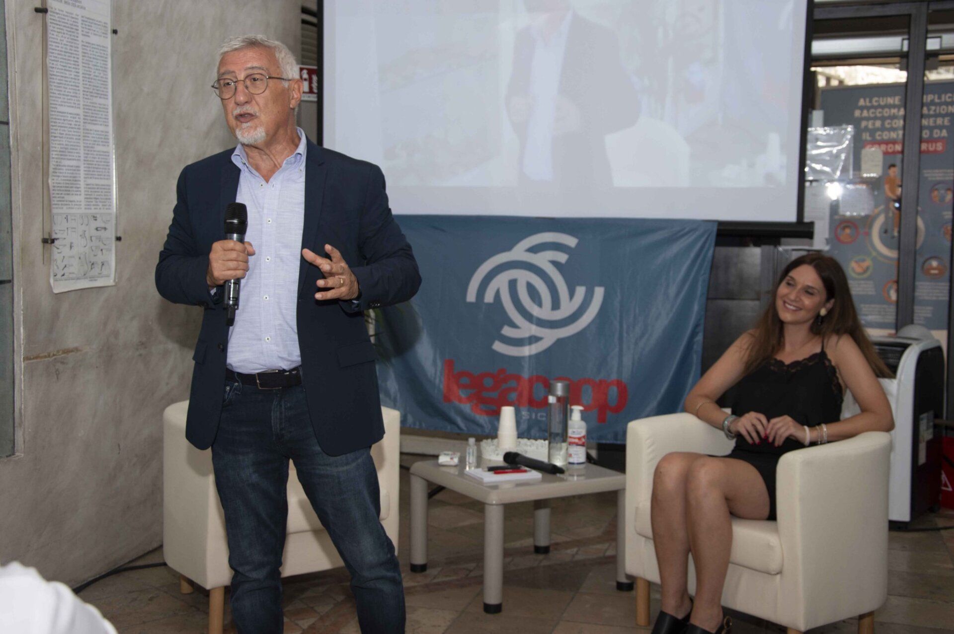 Un uomo è in piedi davanti allo schermo di un proiettore mentre una donna è seduta su una sedia.