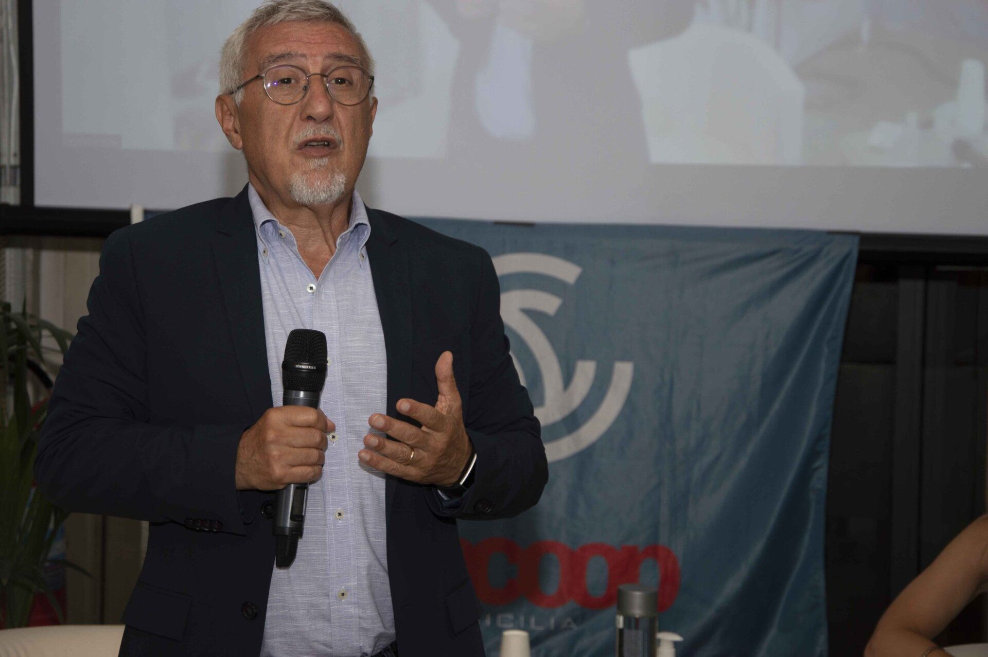 Un uomo in giacca e cravatta tiene in mano un microfono davanti a una bandiera.