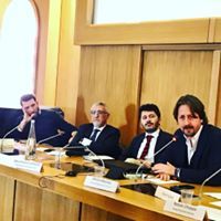 Un gruppo di uomini è seduto a un lungo tavolo con dei microfoni.