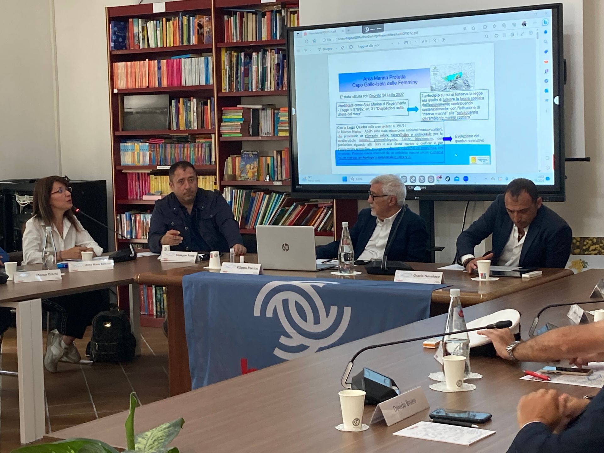 Un gruppo di persone è seduto a un tavolo davanti a un grande schermo.