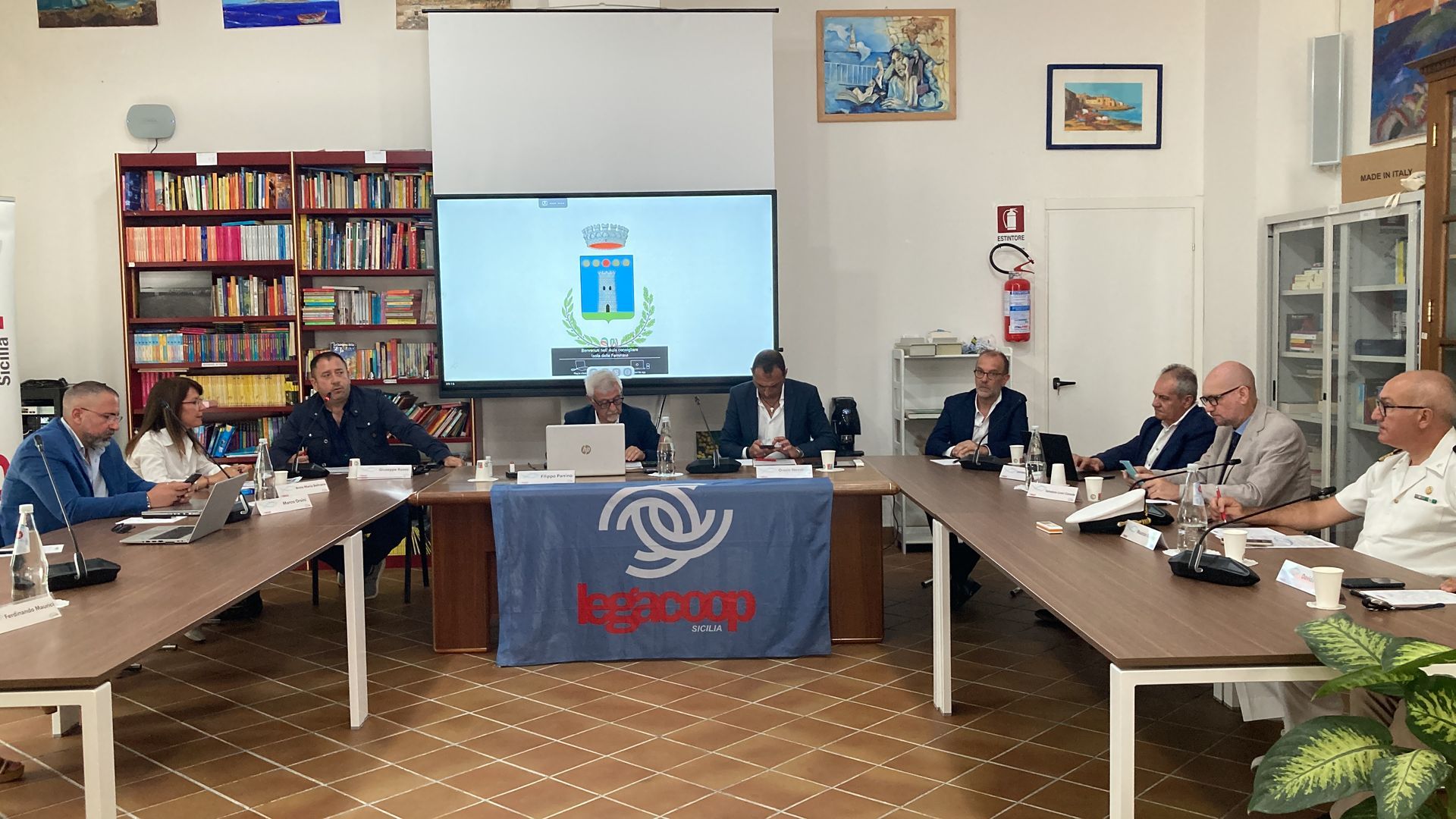 Un gruppo di uomini è seduto attorno a un lungo tavolo davanti allo schermo di un proiettore.