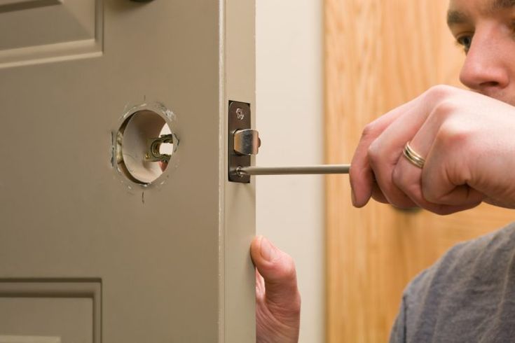 Person installing a door latch with a screwdriver on a light beige door in a wooden frame.