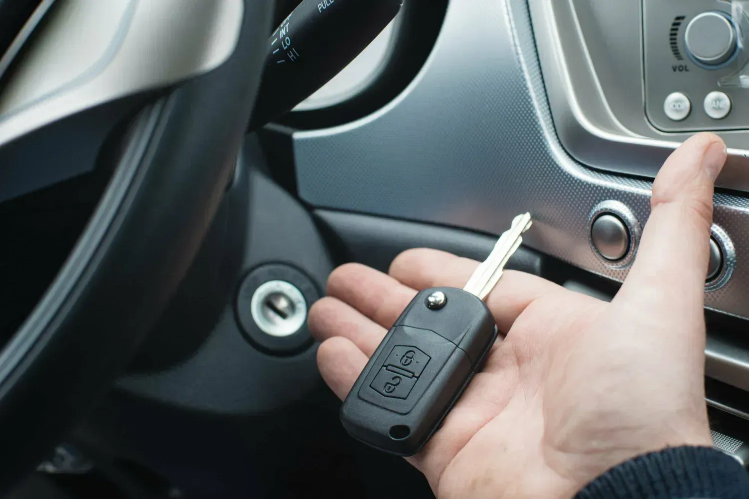 Hand holding a car key inside a vehicle, next to the ignition and dashboard buttons.
