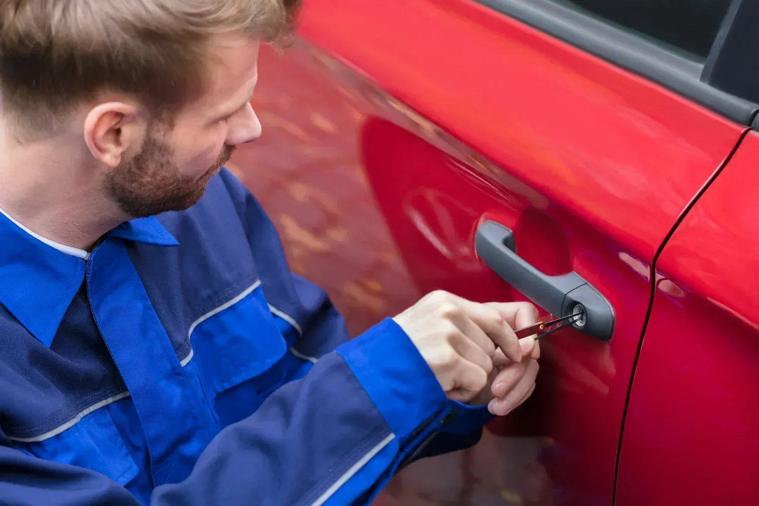 Man in blue jumpsuit using a tool to unlock the door of a red car.