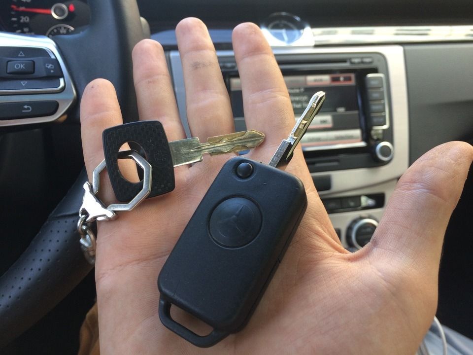 Hand holding car keys: a black fob, two keys, and a black ring. Interior car setting.