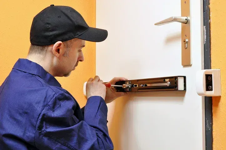 A person in a blue jumpsuit installs a door closer with a screwdriver on a white door.