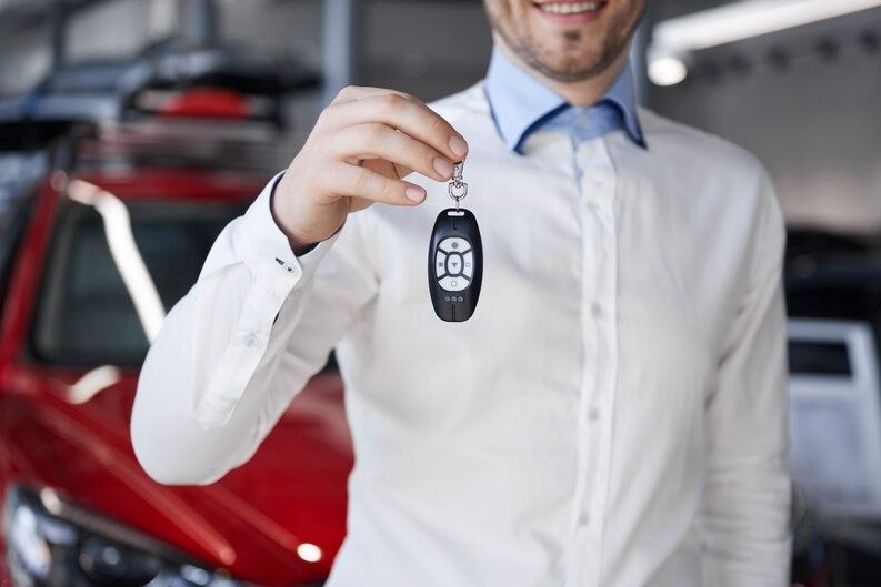 Man in white shirt smiles, holding car key fob in front of a red car at a dealership.