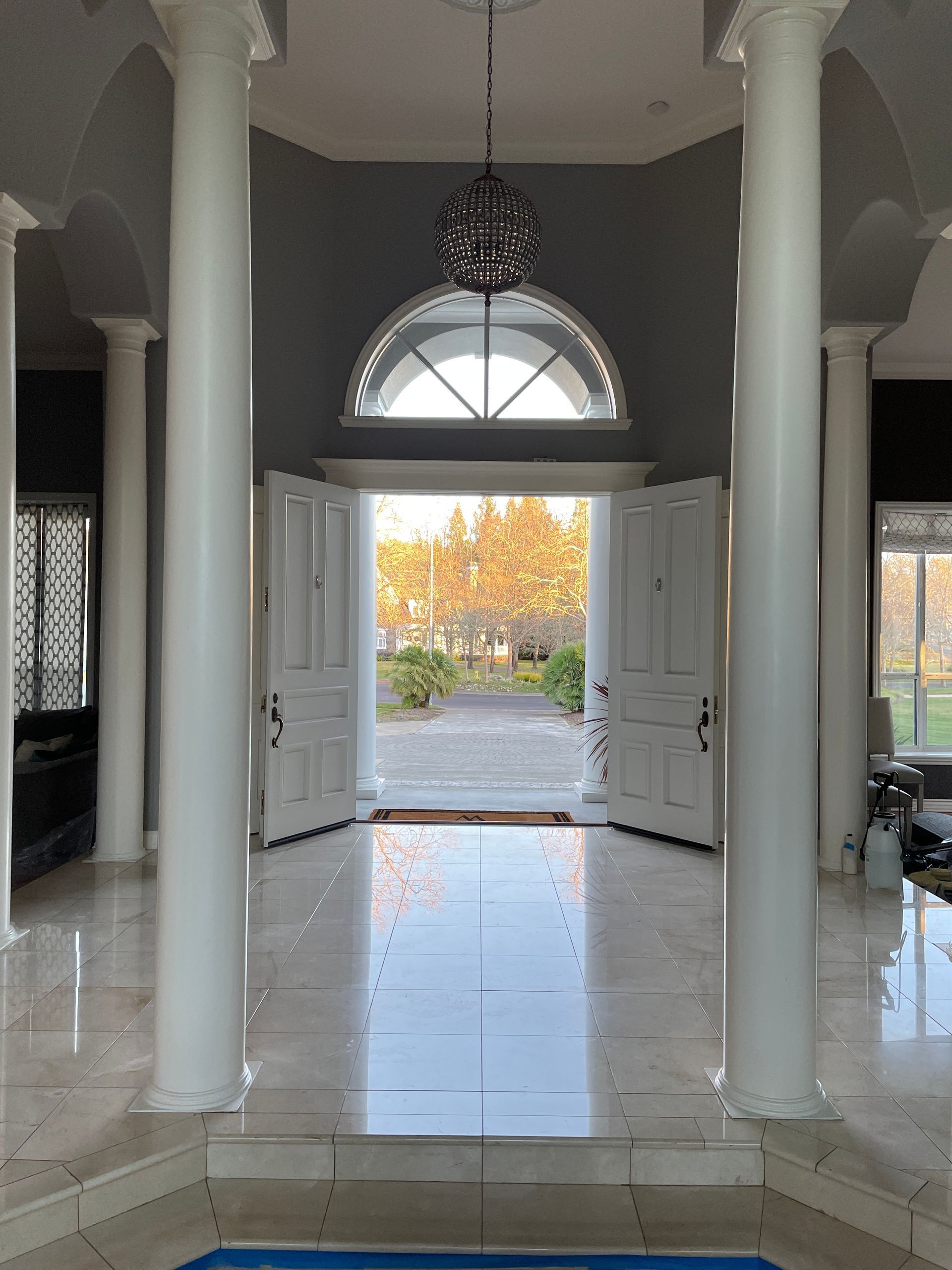 A marble-floored entryway with two white columns leading to an open doorway revealing a sunlit, tree-lined outdoor path.