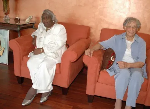 Two people seated on orange armchairs in a living room, one holding a red handbag.