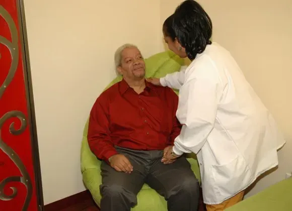 Caregiver in white helps seated patient in red shirt on a green chair in a clinic room