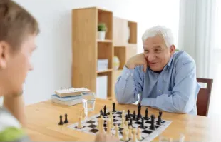 Two people playing chess at a wooden table, with one smiling and resting his head on his hand.