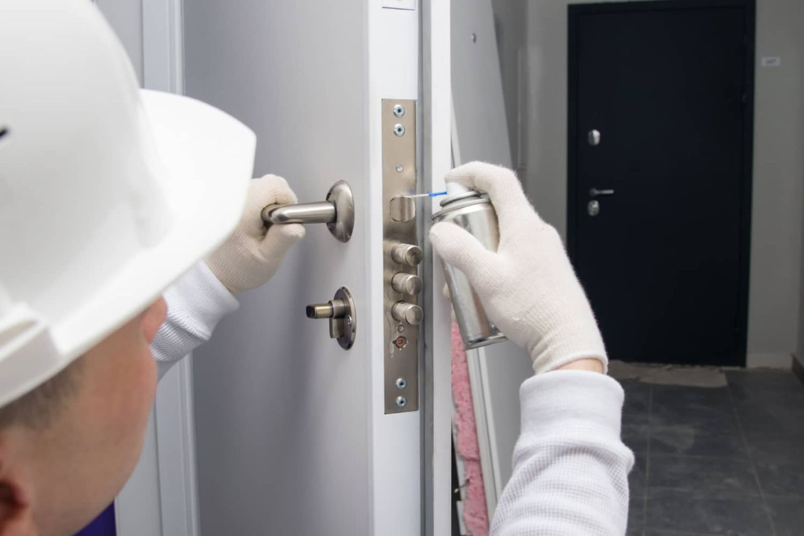 Person in hard hat and gloves spraying lubricant on a door lock.