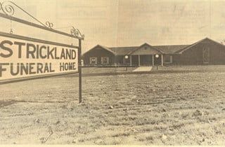 Strickland Funeral Home sign in front of a single-story building on a grassy, unpaved lot.