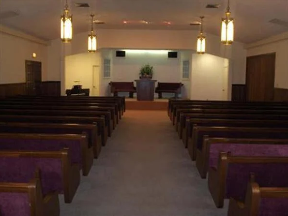 An empty chapel with rows of pews, a stage with a podium and flowers, and hanging lights.