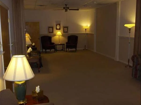 An empty beige room with lamps, chairs, and framed pictures; likely a waiting or sitting area.