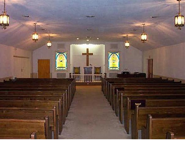 Interior view of a church sanctuary with rows of wooden pews, a cross, and stained-glass windows.