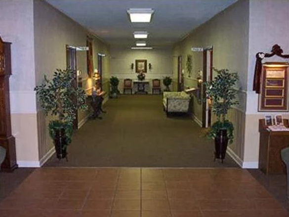 Long hallway in a building with doors, plants, and furniture; brown and beige color scheme.