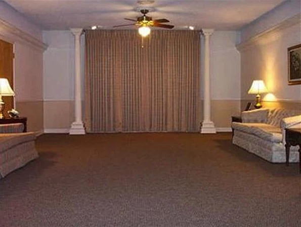 Empty room with beige walls, brown carpet, beige curtains, two couches and a ceiling fan.