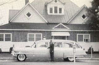 Man standing by a vintage hearse in front of a house. Black and white photo.