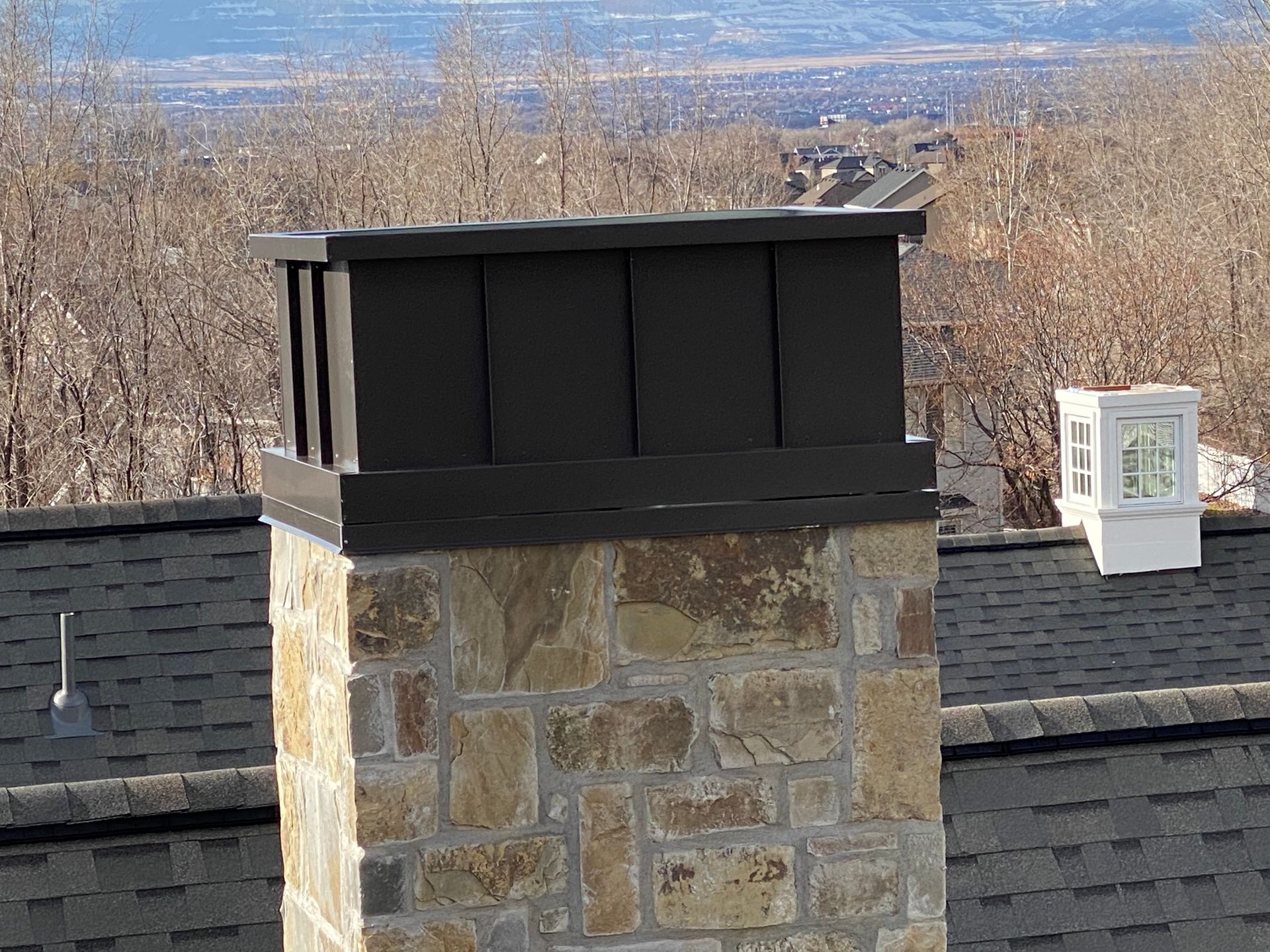 Dark metal chimney cap atop a stone chimney on a rooftop with a cityscape in the background.