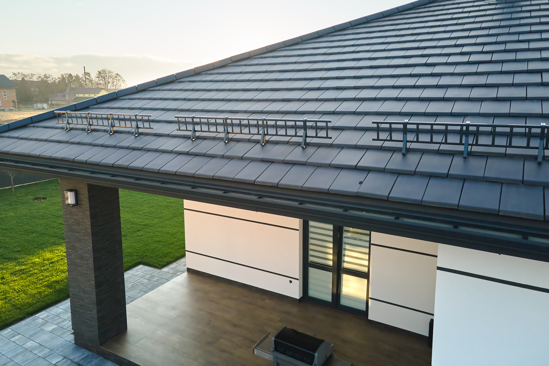 View of a modern home with dark roof tiles, black roof ladders, and a covered patio.