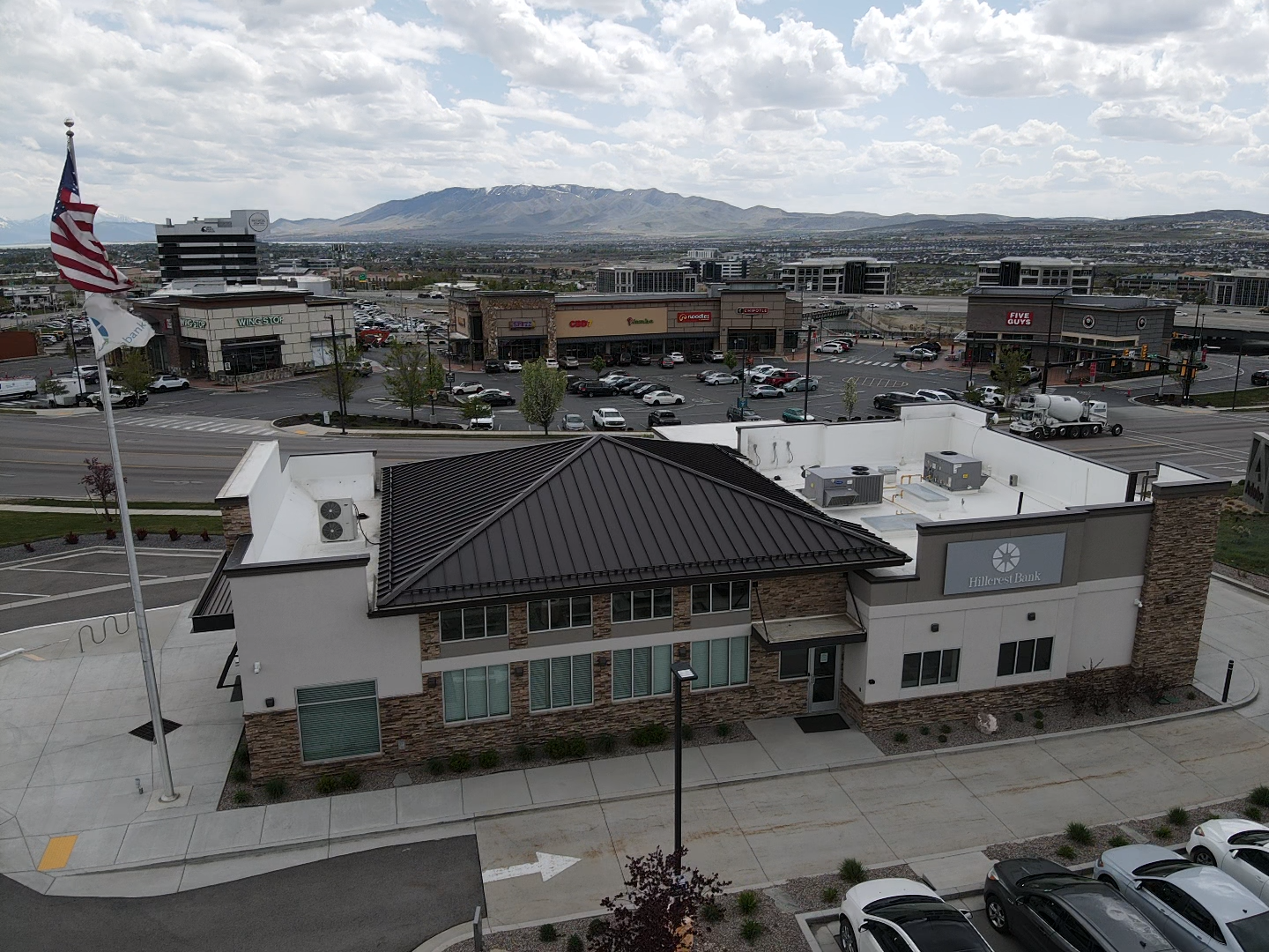 Building with an American flag, other buildings, parking lot, and mountain in the background under a cloudy sky.