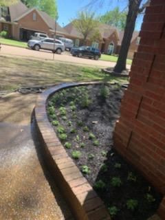 Brick-bordered flower bed next to brick house, with freshly planted greenery and street in background.