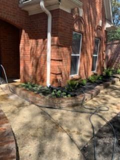 Brick building corner with gutter, windows, and a garden bed. Concrete path with hose on ground.