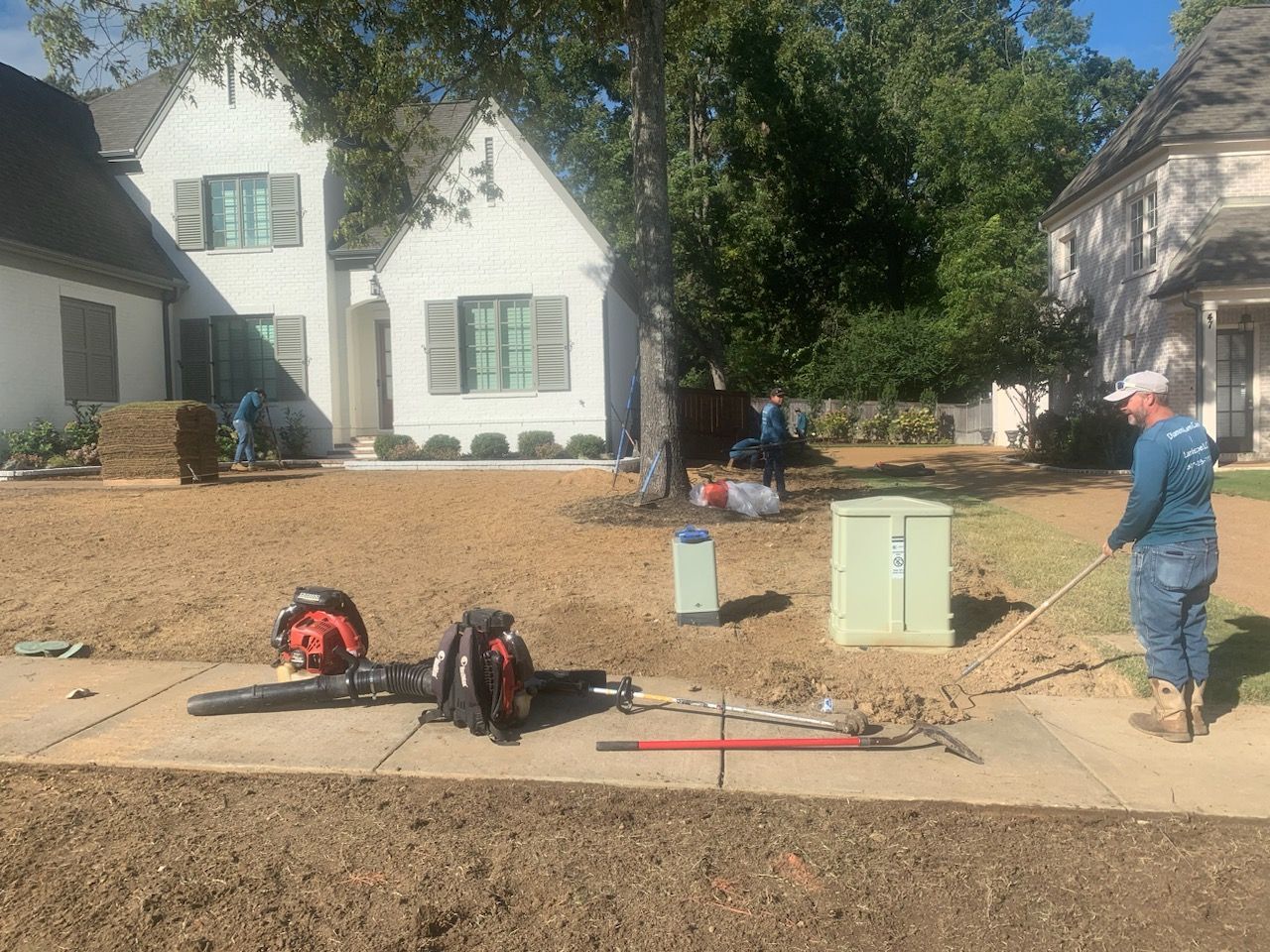 Workers prepping lawn with tools in front of white houses; raking, blowing, and working on dirt.