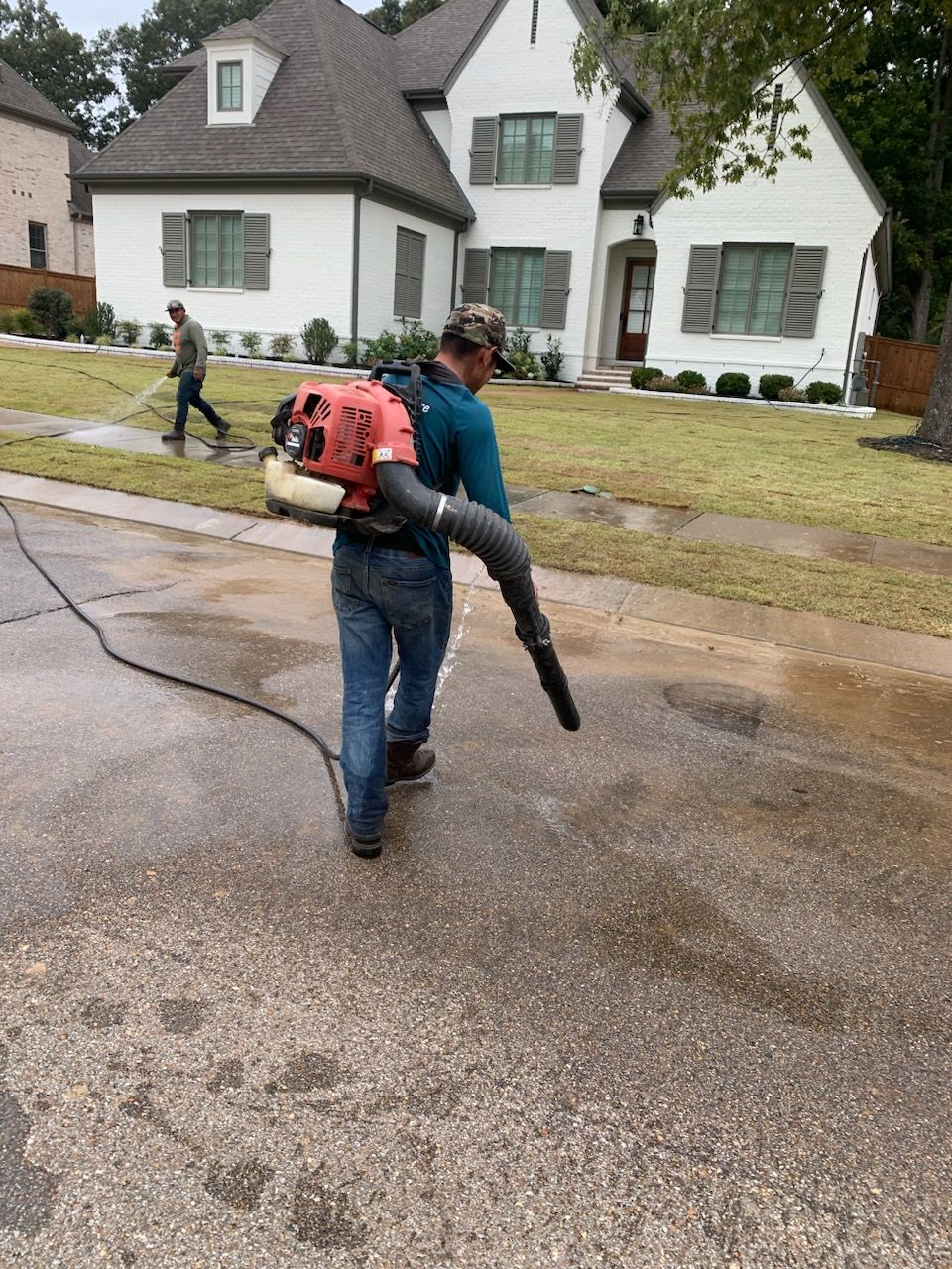 Man in jeans and work boots using a leaf blower on a wet driveway in front of a white house.