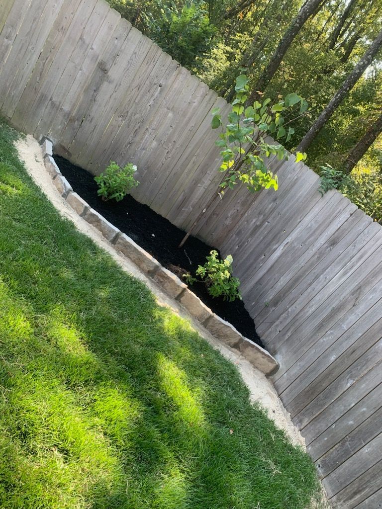 A raised garden bed with dark mulch and small plants, bordered by stones, next to a wooden fence.