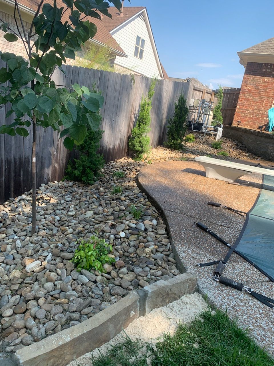 Backyard with rock garden, wooden fence, and path leading to a pool. Sunny day.