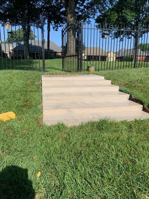 Concrete steps leading up to a wrought iron gate in a grassy yard. Homes visible in the background.