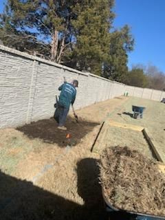 Man spreading mulch in a yard near a stone wall on a sunny day.
