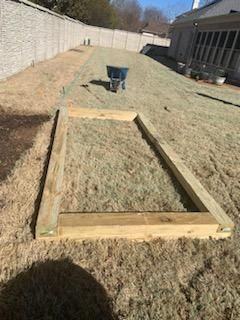 Wooden frame on a grassy hill, likely for a garden bed, wheelbarrow in the background, sunny day.