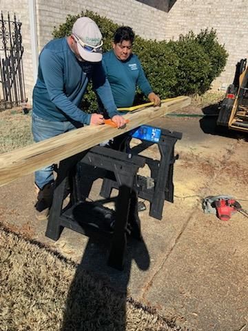 Two men measuring a wooden plank on sawhorses outdoors, one marking with a pencil.