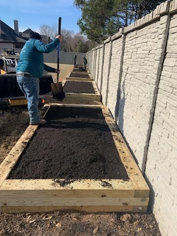 Man shovels soil into wooden garden beds against a textured concrete fence.
