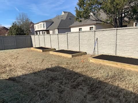 Three raised garden beds in a yard, bordered by a concrete fence and houses under a blue sky.