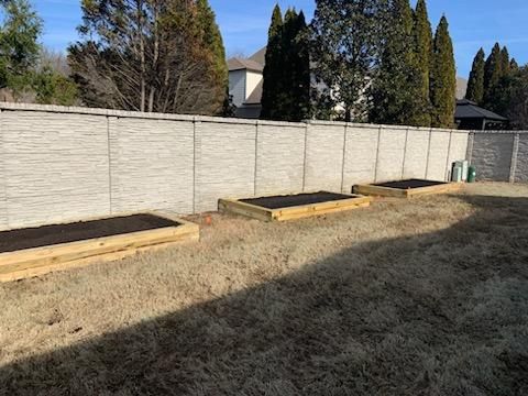 Three wooden raised garden beds on a grassy lawn, with a textured concrete fence in the background.