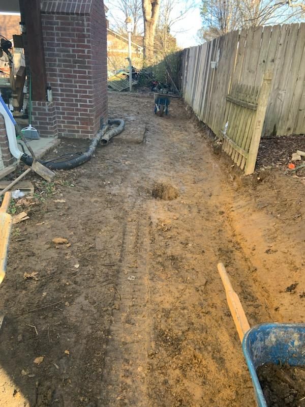 Dirt path along a fence and brick wall. Wheelbarrow and debris present, likely construction area.