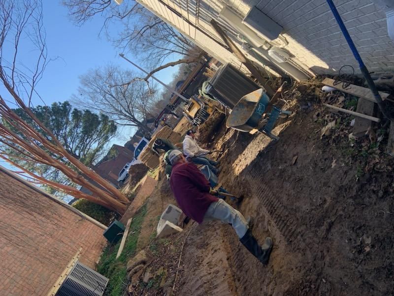 Person digging in dirt alongside a building. Brick wall, green grass, and equipment are visible.