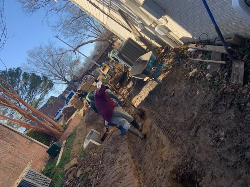 Person in work clothes and mask outside near a house, operating a wheelbarrow and digging in the dirt.