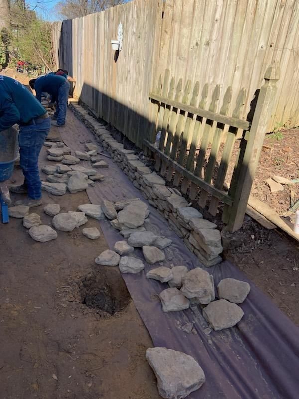 Workers building a stone retaining wall along a wooden fence. Rocks lie on landscaping fabric; dirt ground.