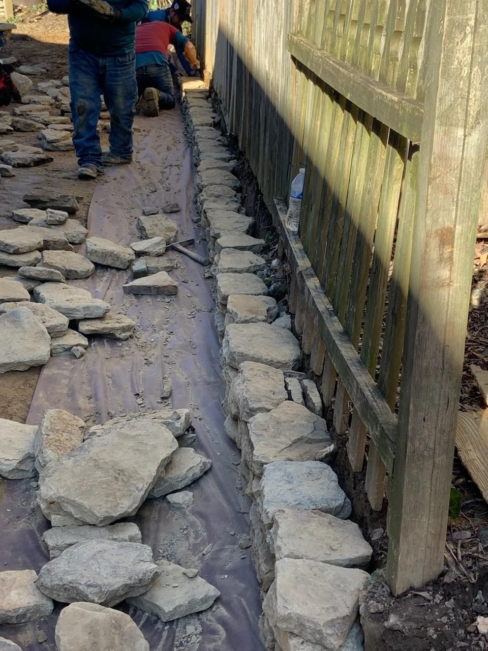 Stone wall being built next to a wooden fence; workers in the background.