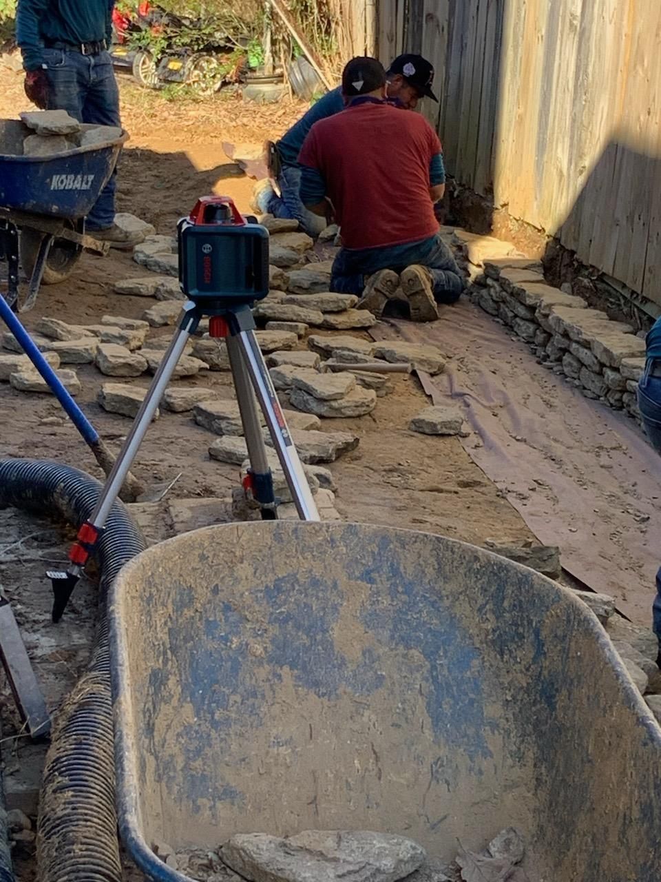 Construction workers laying stone pavers, using a laser level and a wheelbarrow on a dirt path next to a wooden fence.