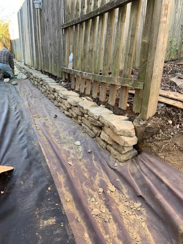 Stone retaining wall built along a wooden fence, with a person working in the background.